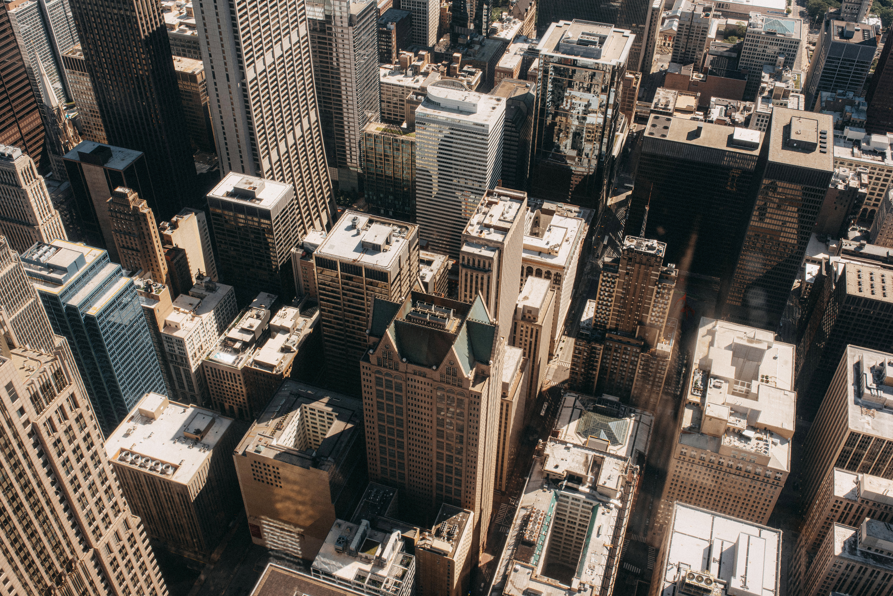 Tall buildings cluster closely in an urban downtown, casting long shadows over the streets below. Various architectural styles and heights create a dynamic cityscape under clear daylight.